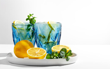 Two blue transparent glasses with cold lemonade stand on a white table, lemons, ice and mint lie next to it, selective focus on a lemon slice, close-up, copy space