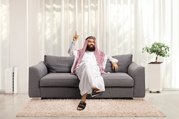 Fototapeta premium Arab man sitting on a gray sofa in a living room at home and pointing up