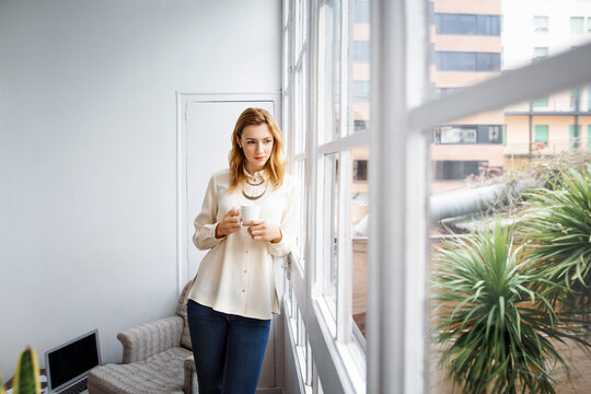 Thoughtful businesswoman leaning on window while holding coffee cup in office