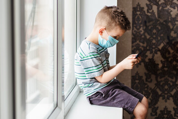 A cute child in a medical mask sits at home in quarantine by the window with a phone in his hands and looks into it. Prevention of coronavirus and Covid - 19