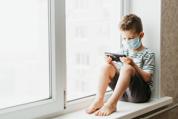 A cute child in a medical mask sits at home in quarantine by the window with a phone in his hands and looks into it. Prevention of coronavirus and Covid - 19