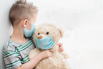 a small child in a mask sits at home in quarantine and looks out the window in a place with a teddy bear. Prevention of Coronavirus and Covid - 19. Concept