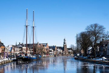 Fototapeta na wymiar People skate on the ice in Lemmer with a ketch frozen in the ice on the left and the reformed church at the back. Sunny winter scene 