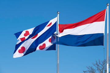 Waving Dutch and Frisian flag. The Frisian flag has four cobalt blue and three white diagonal stripes. The white strips have red pompoms. The Dutch flag is a horizontal tricolor in red, white and blue