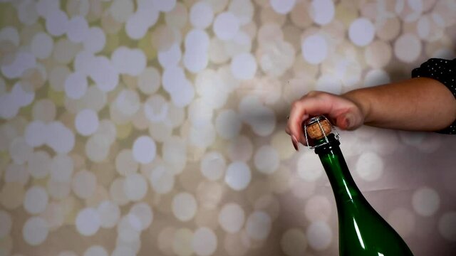 A champagne bottle being opened and popping out and flying across the room, opened by a woman with long glittery nails on a glittery background