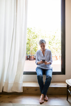 Full length of mature woman using smart phone while sitting on window sill at home