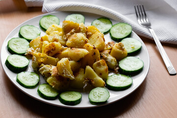 Fried potato slices in a beaten egg with protein, around a circle of green juicy fresh cucumber. Dish on a white plate on a wooden table
