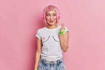 Pretty smiling girl with bob pink hair wears wireless headphones on ears makes korean like sign...