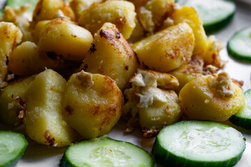 Fried potato slices in a beaten egg with protein, around a circle of green juicy fresh cucumber. Dish on a white plate on a wooden table