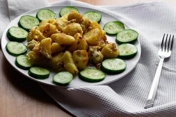 Fried potato slices in a beaten egg with protein, around a circle of green juicy fresh cucumber. Dish on a white plate on a wooden table