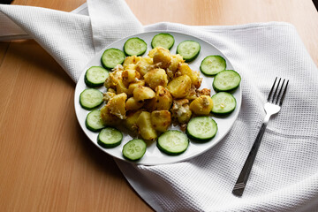 Fried potato slices in a beaten egg with protein, around a circle of green juicy fresh cucumber. Dish on a white plate on a wooden table