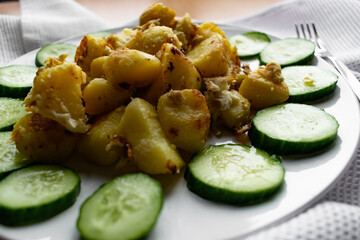 Fried potato slices in a beaten egg with protein, around a circle of green juicy fresh cucumber. Dish on a white plate on a wooden table