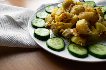 Fried potato slices in a beaten egg with protein, around a circle of green juicy fresh cucumber. Dish on a white plate on a wooden table