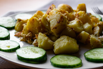Fried potato slices in a beaten egg with protein, around a circle of green juicy fresh cucumber. Dish on a white plate on a wooden table