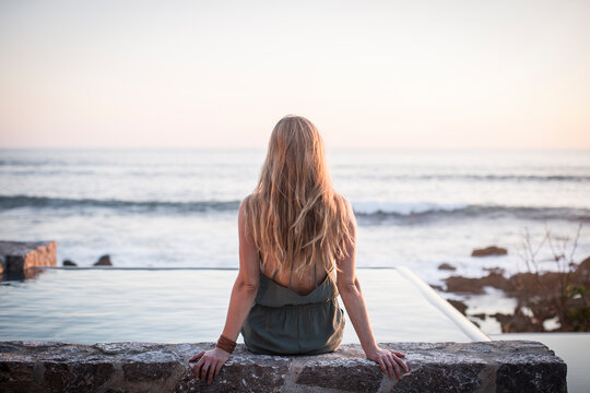 Rear view of woman sitting on retaining wall at artificial pond against sea and clear sky