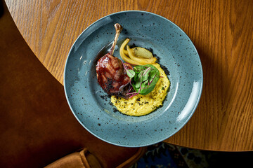 French cuisine - duck leg confit with polenta and caramelized pear, served in a blue plate on a wooden background. Restaurant food. View from above