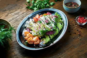 Delicious and dietary salad with grilled shrimp, feta cheese, tomatoes and avocado served in a black plate on a wooden background. Restaurant food