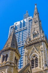 A modern office building towers over a gothic stone church on a blue sky day, in the city of Melbourne, Australia.