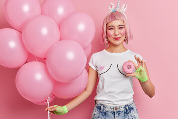 Pleased pink haired girl dressed in t shirt jeans holds sweet tasty donut enjoys party time holds bunch of inflated balloons celebrates special occasion poses indoor. People and holiday concept