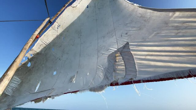 Old traditional maritime traditional vessel Dhow boat sailing under torned sail in the open Indian ocean near Zanzibar Tanzania torn sail bottom view. Traveling 4K footage concept.