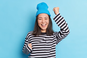 Isolated shot of cheerful happy Asian girl raises hands dances carefree celebrates success smiles broadly expresses sincere emotions and happines wears hat and striped jumper isolated on blue wall