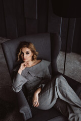 Beautiful young businesswoman in a stylish gray suit sitting on a chair in an office with a modern interior, looking at the camera. Portrait of a charming business woman. Soft selective focus.