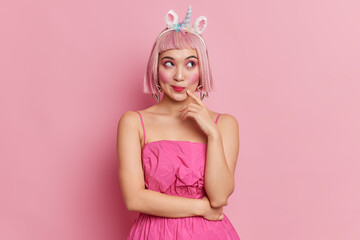 Obraz na płótnie Canvas Studio shot of thoughtful beautiful woman with pink hair looks aside dressed in stylish dress unicorn headband poses against rosy background. Pretty Asian female model thinks about future plans