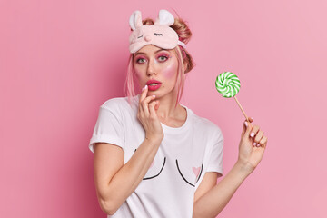 Horizontal shot of serious beautiful woman wears professional makeup looks mysteriously at camera dressed in casual white t shirt soft sleepmask holds lollipop isolated over pink background.