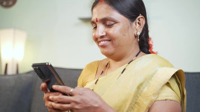 Indian woman busy using mobile phone while sitting on sofa - Concept of people using social media, online messaging app, Internet and entertainment lifestyle