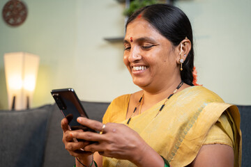 Happy smiling Indian woman busy using mobile phone while sitting on sofa - Concept of people using social media, online messaging app, Internet and entertainment lifestyle.