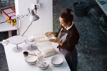 High angle image of a woman examening a bowl under a lamp behind a table with newly produced blank tableware and vases. In a pottery workshop.