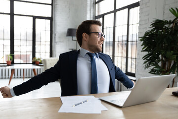 Young male CEO or employer sit at table at workplace stretch after long hours computer work. Caucasian businessman in suit feel excited relieved after job finish, relax rest at desk in office.