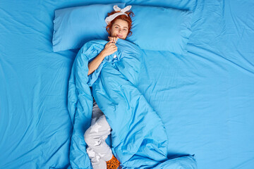 Top view of redhead young woman wrapped in blanket smiles pleasantly looks directly at camera...