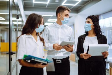 Three business colleagues in protective face mask discussing work related matters on an office building hallway. Teamwork during pandemic in quarantine city. Covid-19.