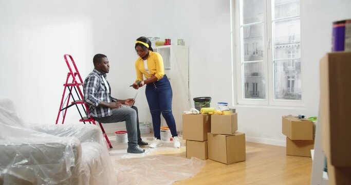 Young wife and husband African American couple in new apartment with boxes during renovation process choosing new wall paint color and typing on laptop searching internet, choosing room design