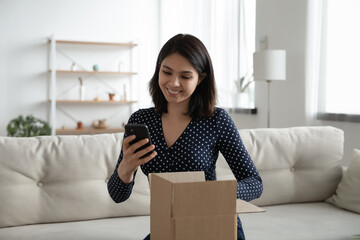 Smiling woman satisfied client hold opened cardboard box parcel text positive feedback to shop website. Grateful young asian female call friend on phone to thank for birthday surprise received by mail