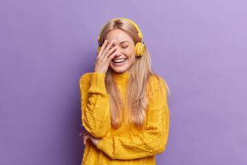 Overjoyed young European woman with blonde hair laughs out loudly makes face palm listens music via wireless headphones wears casual yellow sweater isolated over purple background. Hobby concept