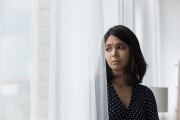 Sad upset young asian woman stand by window hide behind curtain look aside think of heartbreak loneliness. Frustrated teenage female having problems suffer of misunderstanding depression. Copy space