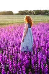 A young beautiful woman with red hair walks in a field with purple flowers in the rays of the setting sun. Active rest, nature walks, digital detox, unity with nature, alone with yourself.