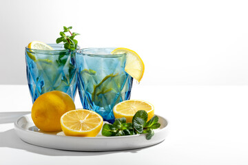 Two beautiful blue glasses with homemade lemonade stand on a white table, lemons and mint lie next to it, selective focus on a lemon slice, close-up, copy space