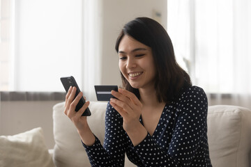 Smiling asian woman capable ebanking app user hold credit card smartphone satisfied with easy quick safe way to pay. Positive young lady sit on couch check bank account balance provide distant payment