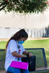 college girl with mask sitting on a bench looking in her briefcase with grass in the background