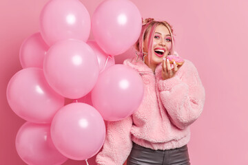 Obraz na płótnie Canvas Positive young teenage girl with bright pink makeup bites delicious sweet doughnut dressed in coat going on birthday party holds inflated balloons isolated over rosy background. Festive concept