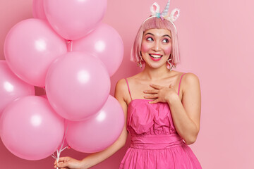 Obraz na płótnie Canvas Horizontal shot of pretty cheerful birthday girl accepts congratulations smiles pleasantly holds helium balloons wears dress has pink hair poses indoor. People festivity special occasion concept