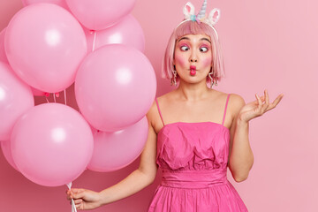 Obraz na płótnie Canvas Beautiful young Asian woman makes fish lips has funny grimace raises hand wears bright makeup raises hand holds helium balloons isolated over pink background celebrates anniversary or birthday