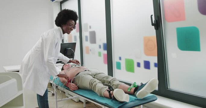 Portrait Of Attractive Positive Afro-American Woman Doctor Looking At Camera. Profession, Hospital, Healthcare. Physician Cardiologist Connects The Patient To The ECG Machine. Preparing To Cardiogram
