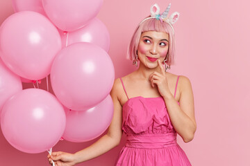 Obraz na płótnie Canvas Photo of thoughtful dreamy young Asian woman wears pink dress hair wig looks aside happily thinks about coming party and celebration holds balloons celebrates first day at work stands indoor