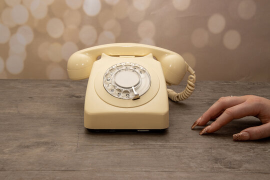 A elegant woman's hand next to an old vintage 70's and 80's style rotary telephone receiver on a wooden background, retro office phone call concept
