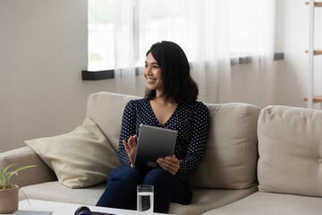 Smiling young asian female sit on cozy couch at home working on electronic tablet. Millennial mixed...