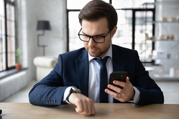 Focused millennial Caucasian male employee in suit look at hand watch clock checking time meet deadline. Serious young businessman hold use modern smartphone, synchronize gadget with smartwatch.